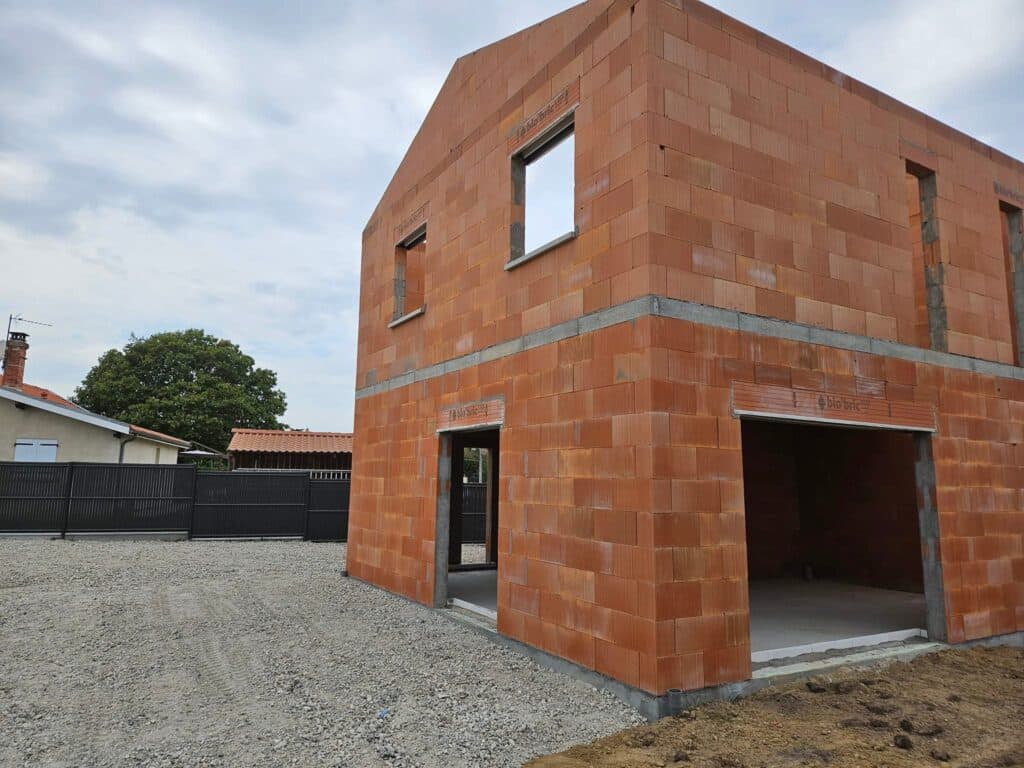 Gros œuvre maison : Avancement avec briques bio'bric. Construction de maison individuelle. Façade en briques terre cuite "bio'bric" sous un ciel nuageux. Gros œuvre achevé avec portes et fenêtres.